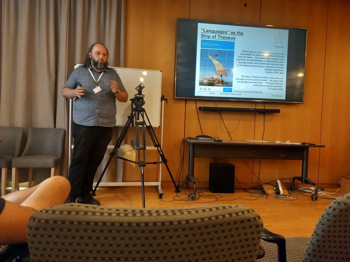 George hält seinen Vortrag auf der Konferenz. Er steht auf einem Podium neben einem Bildschirm und präsentiert eine Powerpoint-Präsentation. Auf der Folie steht "Sprachen" als das Schiff des Theseus".